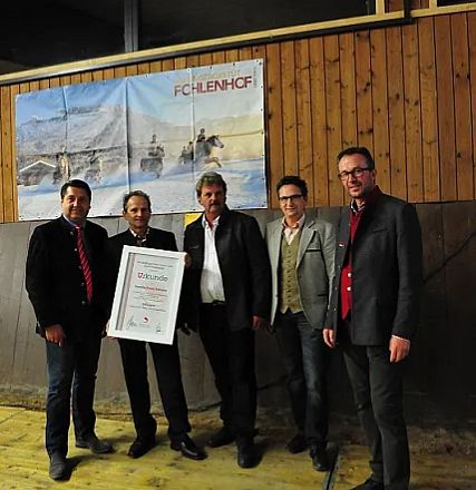 Cinque uomini in abiti formali posano con un certificato incorniciato, davanti a un poster in un ambiente rustico, di legno.