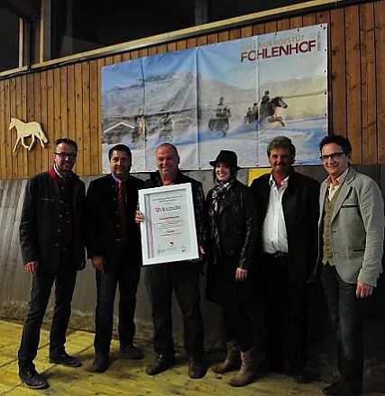 Gruppo di sei persone in piedi con una tela sullo sfondo. Una persona tiene un certificato. L'atmosfera sembra formale e celebrativa.
