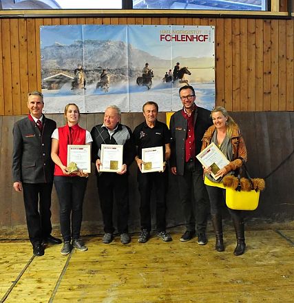 Gruppo di persone sorridenti in un ambiente rustico, alcune con certificati in mano e una donna con un secchio giallo contenente peluche a forma di cavallo.