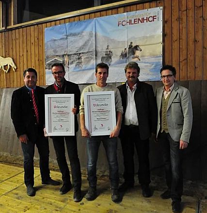 Cinque persone in posa, tre uomini tengono dei certificati incorniciati. Sfondo di legno e una foto di cavalli appesa sopra di loro.