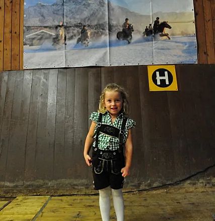 Una bambina in abito tradizionale posa sorridente davanti a un muro in legno con un cartello "H" e uno sfondo di neve e montagne.