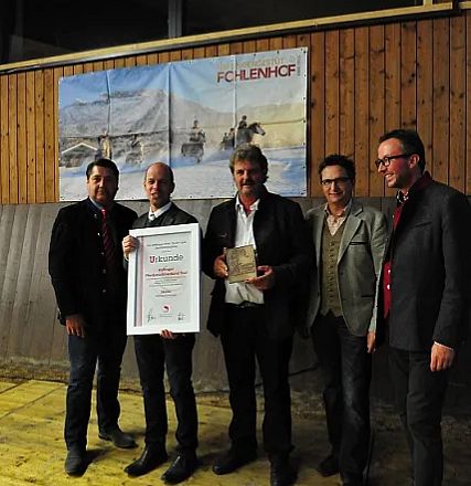 Cinque uomini in abiti formali posano sorridenti. Due di loro tengono premi tra le mani. Sfondo di legno con un grande dipinto appeso.