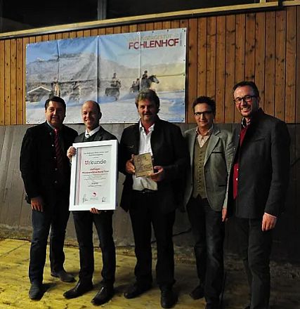 Cinque uomini in abbigliamento formale posano con un certificato e un trofeo, sorridendo davanti a un dipinto di cavalli appeso al muro.