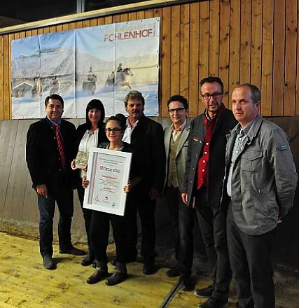 Un gruppo di persone in posa con un certificato, su uno sfondo di legno. Uno di loro tiene un trofeo. Sullo sfondo, un quadro di paesaggio invernale.