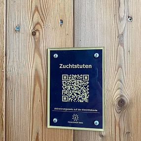A sign on a wooden wall features a QR code and the German word "Zuchtstuten," indicating breeding mares. It has a dark blue background and metallic frame.