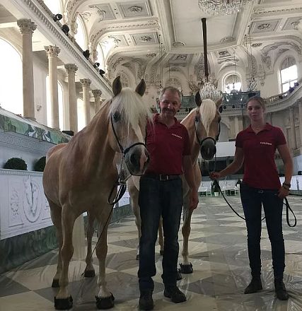 Zwei Personen in roter Kleidung stehen mit zwei blonden Pferden in einer prächtigen, hellen Halle mit verzierten Decken und großen Säulen.