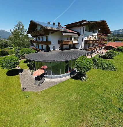 Ein großes, dreistöckiges Gebäude mit Balkonen liegt in einer grünen, ländlichen Umgebung. Der Himmel ist klar und einige Hügel sind im Hintergrund zu sehen.