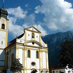 Eine Kirche mit einem hohen Turm und einer Uhr steht vor einem Bergpanorama. Der Himmel ist blau mit einigen Wolken, und Bäume umrahmen das Gebäude.