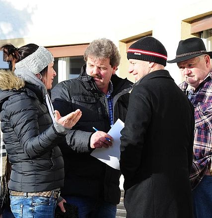 Cinque persone parlano insieme all'aperto, vestite con abiti invernali. Una donna gesticola mentre gli uomini la ascoltano. C'è del sole.
