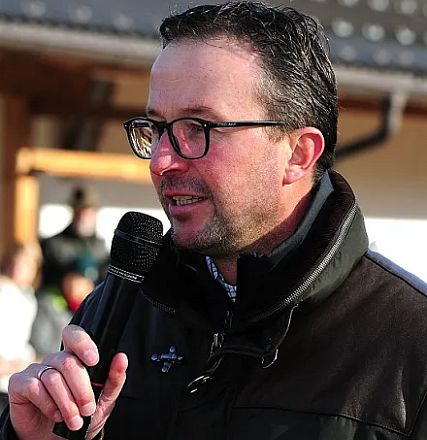 Uomo occhialuto parla al microfono, indossa una giacca nera all'aperto, forse durante un evento o una cerimonia pubblica. Sguardo concentrato.