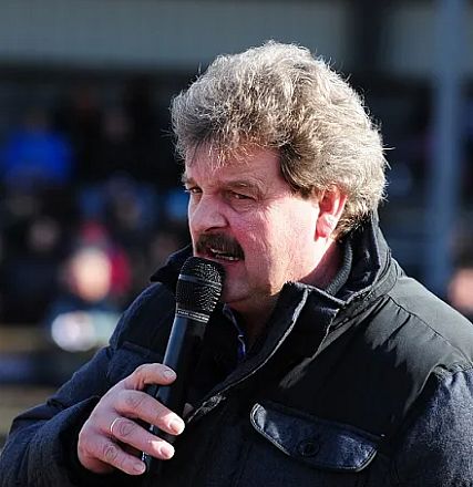 Un uomo parla al microfono all'aperto, indossando una giacca scura. Sullo sfondo, persone sfocate suggerendo un evento pubblico o un'intervista.