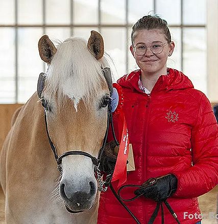 Eine Person in roter Jacke steht neben einem hellen Pferd in einer Reithalle. Beide blicken in die Kamera, im Hintergrund ist eine weitere Person zu sehen.