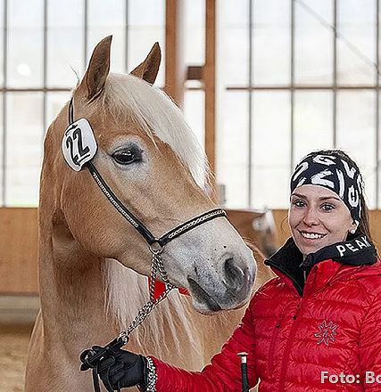 Eine Frau in roter Jacke lächelt neben einem hellbraunen Pferd mit Nummer 22, in einer Reithalle mit Holzkonstruktion und Fenstern im Hintergrund.