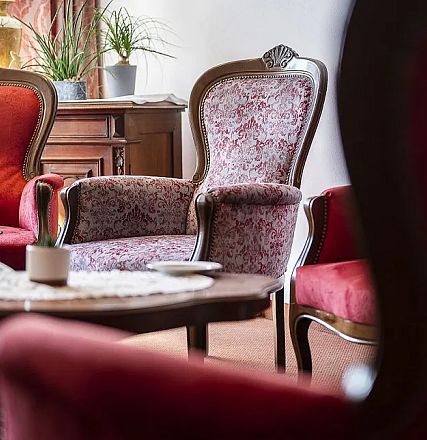 Ein stilvolles Wohnzimmer mit roten und gemusterten Sesseln, einem Holztisch mit Spitzendeckchen und einer Pflanze im Hintergrund.