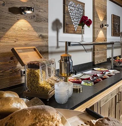 Ein reichhaltiges Frühstücksbuffet in einem gemütlichen Raum. Auf einem Holztresen sind Brot, Müsli, Marmelade und frisches Obst appetitlich präsentiert.