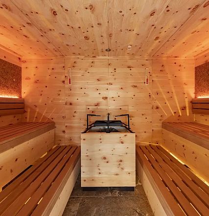 Eine gemütliche Sauna mit Holzwänden und -bänken. Ein Saunaofen steht in der Mitte, warmes Licht schafft eine entspannende Atmosphäre.