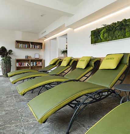 Ein moderner Ruheraum mit sechs grünen Liegen, die in einer Reihe stehen. Die Wände sind weiß, es gibt eine Pflanzendekoration und ein Bücherregal.