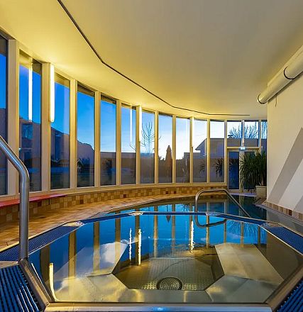 Ein modernes, elegantes Hallenschwimmbad mit großen Fenstern, durch die blaues Abendlicht strömt. Ein Whirlpool ist im Vordergrund zu sehen.