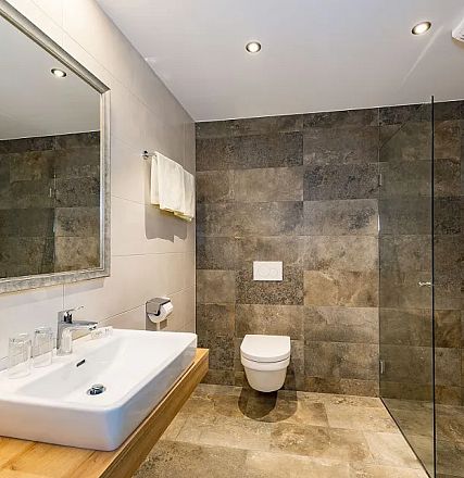 Modernes Badezimmer mit Marmorfliesen, freistehendem Waschbecken auf Holzplatte, wandmontiertem WC und gläserner Duschabtrennung. Elegantes, minimalistisches Design.