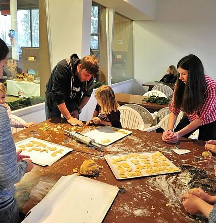 Eine Gruppe von Menschen, darunter Kinder, backt an einem Tisch Plätzchen. Der Tisch ist mit Teig und Mehl bedeckt, alle arbeiten konzentriert.