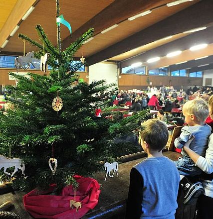 Kinder betrachten einen weihnachtlich geschmückten Tannenbaum in einer großen Halle. Im Hintergrund sind viele Menschen an Tischen versammelt.