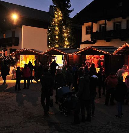 Ein stimmungsvoller Weihnachtsmarkt bei Dämmerung mit leuchtenden Holzbuden, geschmückten Tannenbäumen und Menschen, die die festliche Atmosphäre genießen.