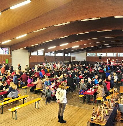 Große Halle mit Holzdecke, in der viele Menschen an Tischen sitzen. Auf der Bühne im Hintergrund scheint eine Aufführung oder Redebeitrag stattzufinden.