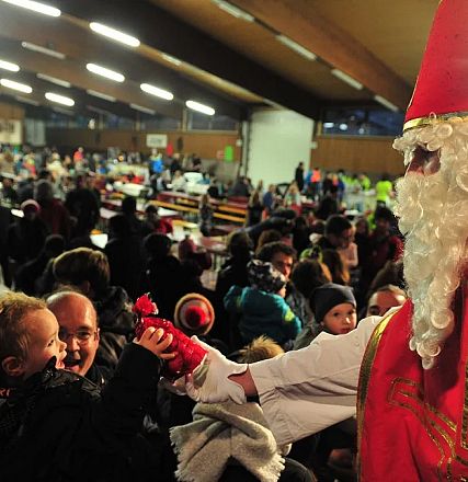 In einer großen Halle überreicht ein als Nikolaus verkleideter Mann ein Geschenk an ein Kind. Viele Menschen sitzen an Tischen im Hintergrund.