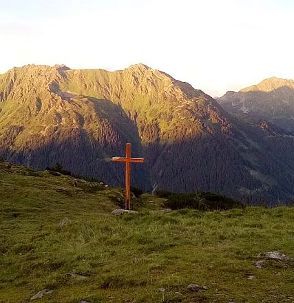 A scenic Alpine landscape at sunset with a wooden cross on a grassy hill, overlooking a majestic mountain range. A horse grazes peacefully in the foreground.