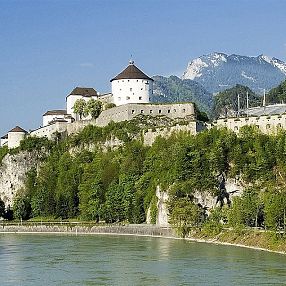 Eine historische Festung thront auf einem Hügel über einem Fluss, umgeben von bewaldeten Bergen und einer klaren, blauen Himmelkulisse.