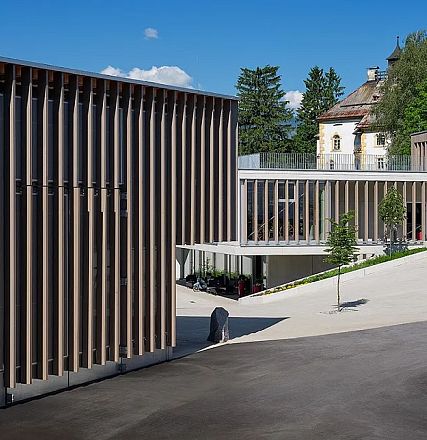 Modern architecture with vertical slats and large windows, surrounded by trees and a clear blue sky. A historic building is visible in the background.