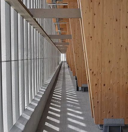 A modern architectural space with angled wooden panels and metal beams casting dynamic shadows along a narrow, sunlit corridor.