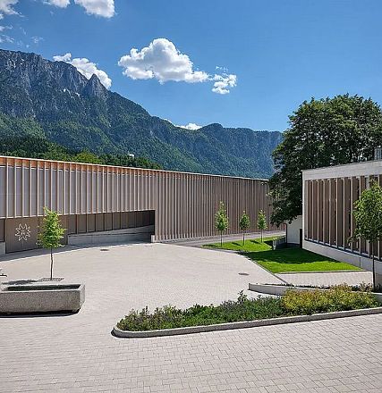Modern building with wooden facade set against a backdrop of mountains and blue sky. Courtyard features a few small trees and a stone seating area.