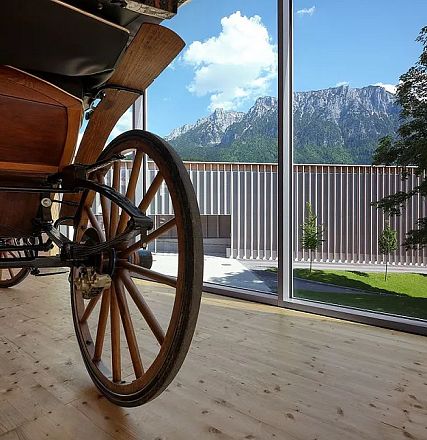 Antique wooden carriage wheel inside a modern building with large windows, offering a view of majestic mountains and a lawn under a clear blue sky.