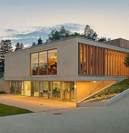 Modern building with large glass windows, surrounded by greenery, at twilight. The structure has wooden panel accents and is set against a serene sky.