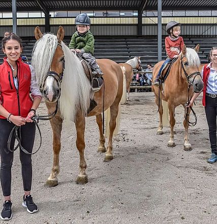 Zwei Kinder in Reitkleidung sitzen auf zwei Pferden in einer Reithalle, begleitet von zwei Erwachsenen in roten Westen. Im Hintergrund sind Tribünen sichtbar.