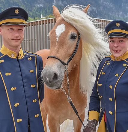 Zwei uniformierte Personen stehen lächelnd neben einem Haflinger mit blonder Mähne. Im Hintergrund sind Berge und ein Gebäude zu sehen.