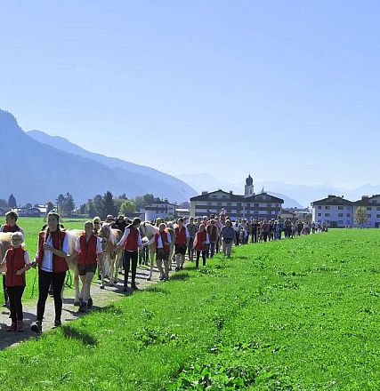 Eine Gruppe von Menschen wandert auf einem Pfad durch eine grüne Wiese mit Bergen im Hintergrund. Ein Reiter führt ein Pferd an der Spitze der Gruppe.