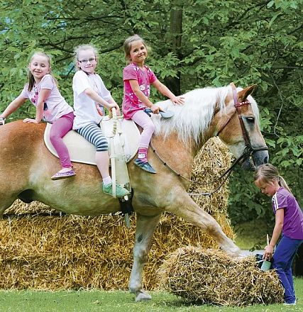 Vier Kinder spielen im Freien auf einem großen Pferd neben Strohballen. Im Hintergrund sind Bäume und im Vordergrund hilft ein Kind beim Balancieren.