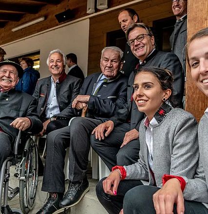 Eine Gruppe von Menschen, teils in Tracht gekleidet, lächelt und sitzt in einem überdachten Bereich. Einige tragen traditionelle Hüte, andere Uniformen.