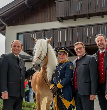 Sechs Personen posieren mit einem hellbraunen Pferd vor einem traditionellen Holzhaus. Die Gruppe ist in festlicher und traditioneller Kleidung gekleidet.