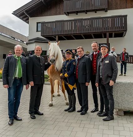 Eine Gruppe von sechs Männern, einige in traditioneller Uniform, posiert neben einem Pferd vor einem modernen Gebäude mit Holzverkleidung.