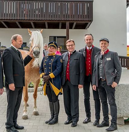 Eine Gruppe von Menschen steht neben einem hellen Pferd vor einem Gebäude. Die Personen tragen formelle und historische Kleidung. Der Himmel wirkt bewölkt.