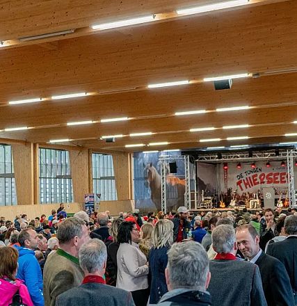 Menschenmenge in einer großen Halle mit Holzdach. Eine Bühne im Hintergrund wird von Musikern bespielt. Viele Besucher stehen und unterhalten sich.