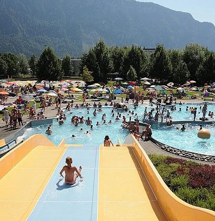 Kinder und Erwachsene genießen einen sonnigen Tag in einem Freibad mit großen Wasserrutschen und zahlreichen Sonnenschirmen vor einer bergigen Landschaft.