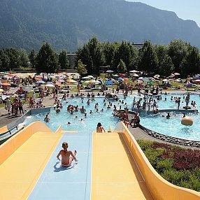 Kinder und Erwachsene genießen einen sonnigen Tag in einem Freibad mit großen Wasserrutschen und zahlreichen Sonnenschirmen vor einer bergigen Landschaft.