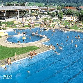 Ein Freibad mit mehreren Becken, vielen Menschen beim Schwimmen und Entspannen. Im Hintergrund sind Gebäude und grüne Hügel sichtbar, sonniges Wetter.