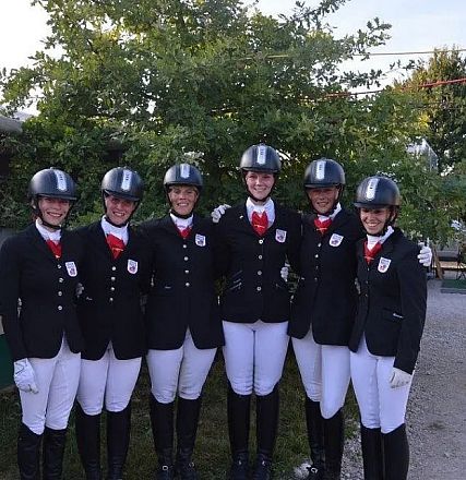 Sechs Reiterinnen in schwarzen Reitjacken und weißen Hosen stehen nebeneinander. Sie tragen Helme und rote Halstücher. Im Hintergrund sind Bäume und Stände.