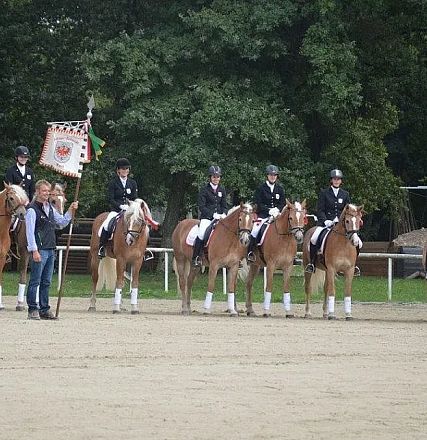 Gruppe von sieben Reitern in Uniform auf Pferden, zwei Personen stehen nebendran, eine hält ein Banner. Sie befinden sich auf einem Sandplatz vor Bäumen.