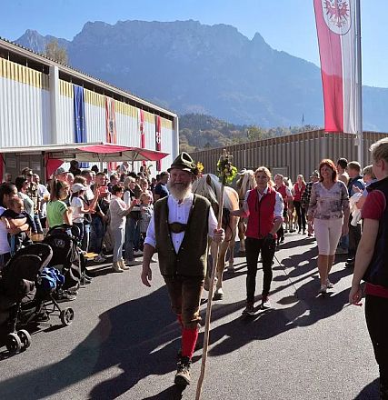 Menschen auf einem sonnigen Festgelände, einige tragen traditionelle Kleidung. Im Hintergrund sind Fahnen und ein beeindruckendes Bergpanorama zu sehen.